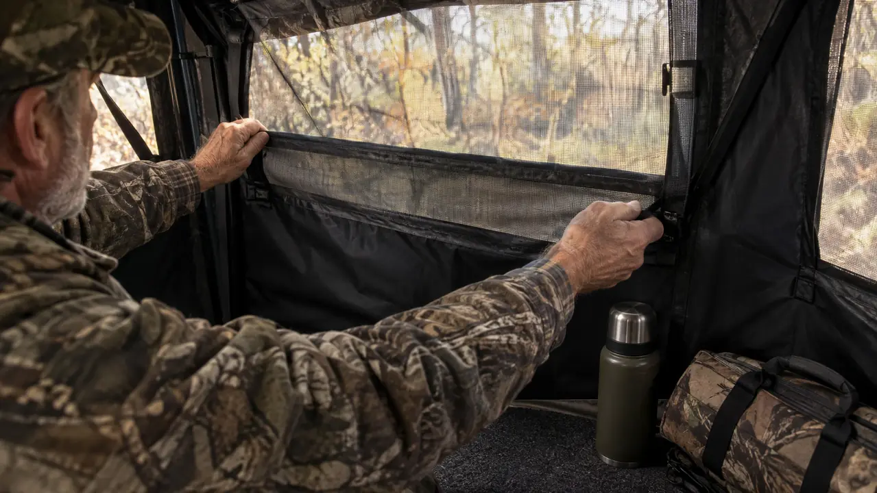 Hands adjusting the mesh window of a hunting blind from inside, with a thermos and carry bag on the floor
